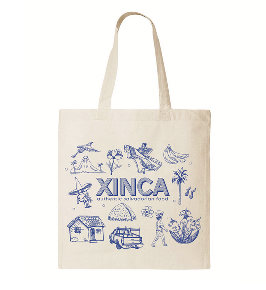 A canvas tote bag with blue design, showing a woman dancing, bananas, flor de izote, a truck, a bird, palm trees and a house with the Xinca authentic Salvadorian food logo in the middle